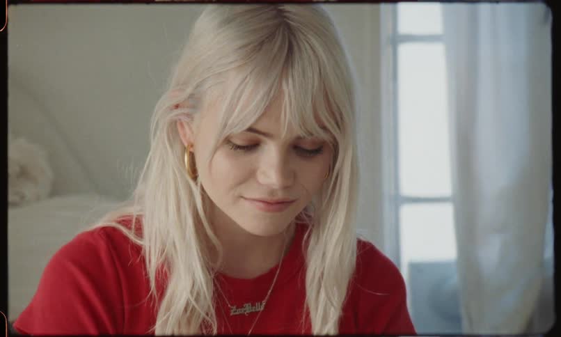 A blonde haired pendant wearing woman sits on a bed and reads a hand written note