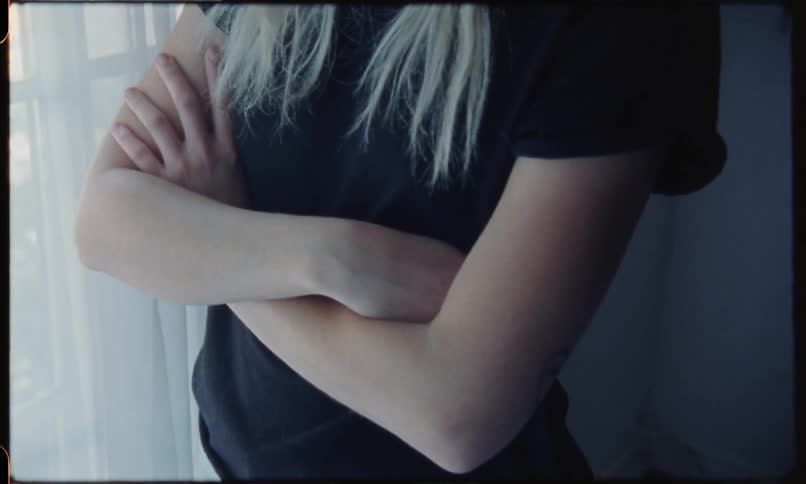 A blonde female has an unlit cigarette in her mount as she stands in a black t shirt by the window