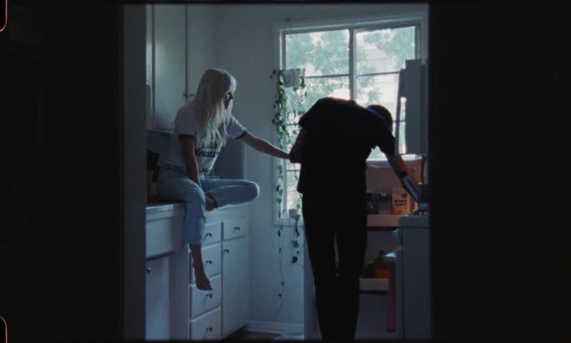 A blonde haired woman sits on a kitchen counter top and talks to a man who opens the fridge