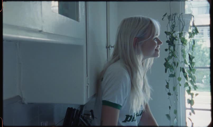 A pale female teenager with long very light blonde hair is leaning against a cupboard in a kitchen talking to someone