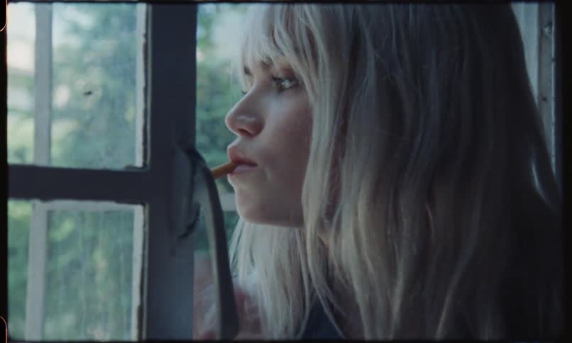 A blonde female sits on a window sill in a denim jacket and lights a cigarette