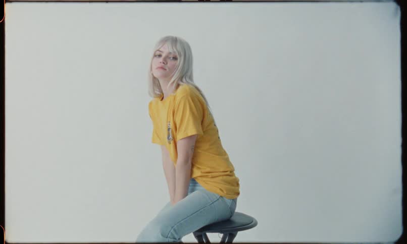 A pale white teenager with light blonde hair is sitting a stool transitioning through various poses during a photo shoot