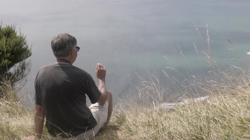 A man sits on the edge of a cliff holding a weed on a cloudy day