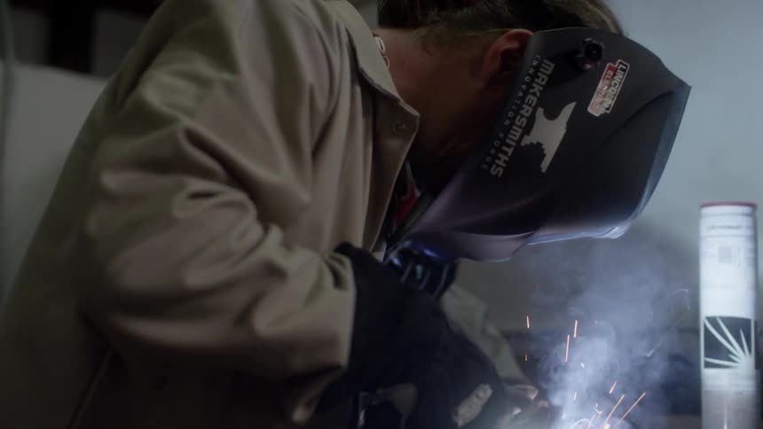 A white man wearing a welding helmet welds an object
