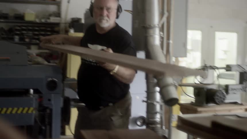 Man with white beard wearing headphones and black tshirt with white logo carries a board from the end of a table saw and places it on a stack