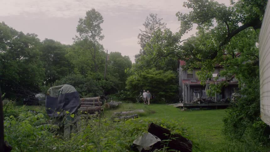 Two men are walking across a cluttered lawn next to a rundown house