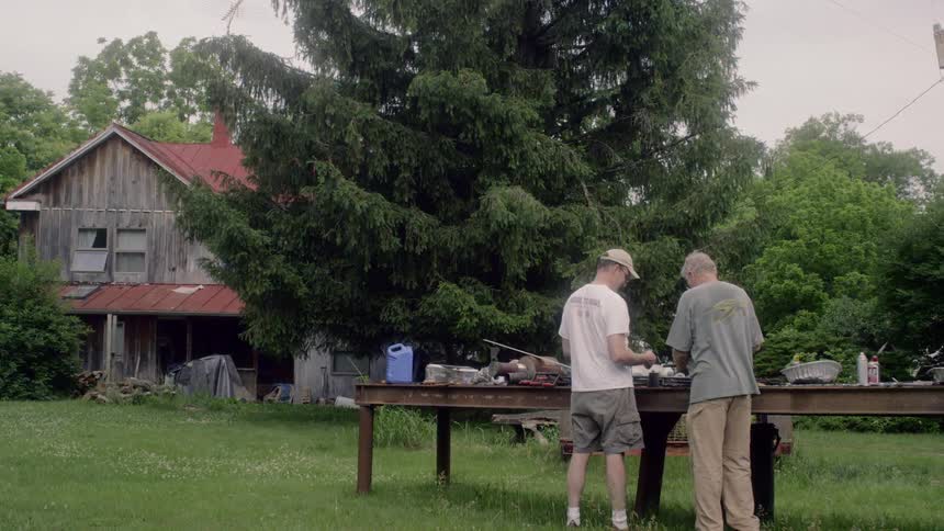 Two men stand at a metal work bench in a yard near a barn