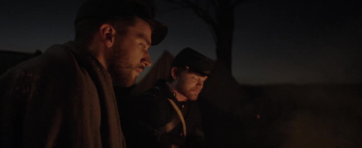 Two young men in old military garb sit by a camp fire at night