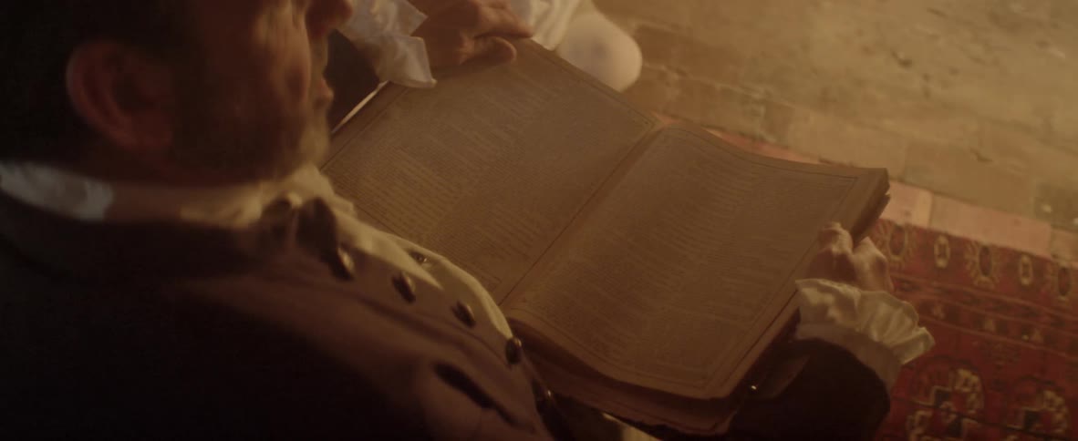 A man dressed in clothing from several centuries ago sits and reads aloud from a large book