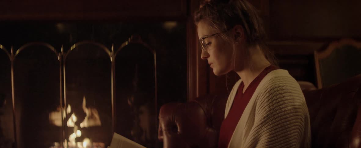 A woman sits in a leather chair while reading a book next to a burning fire in a fireplace