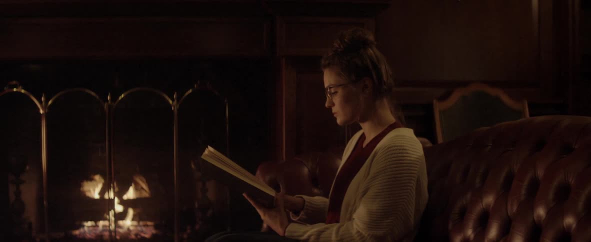 A white female wearing a cardigan sweater and eyeglasses is sitting on a dark red sofa reading a book in front of a fireplace
