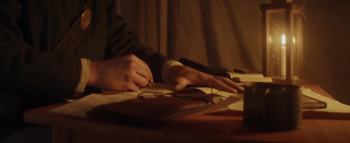 A man sits at a desk and writes by candlelight