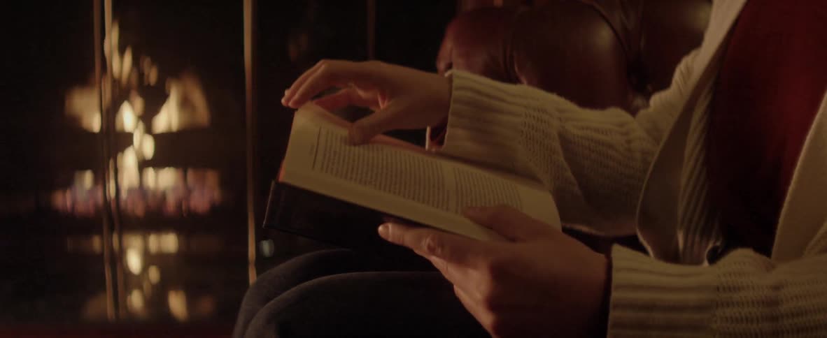 A person wearing a cardigan sits on a leather chair reading a book by an open fire