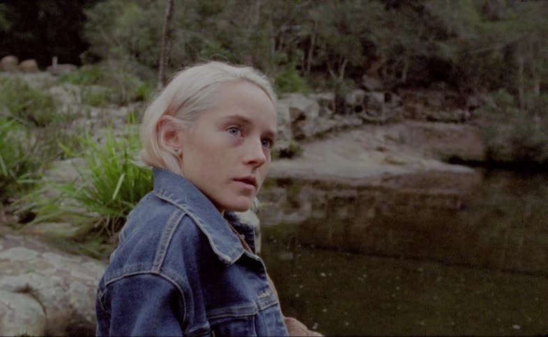 A blonde woman in a denim jacket sits by a lake and turns her head