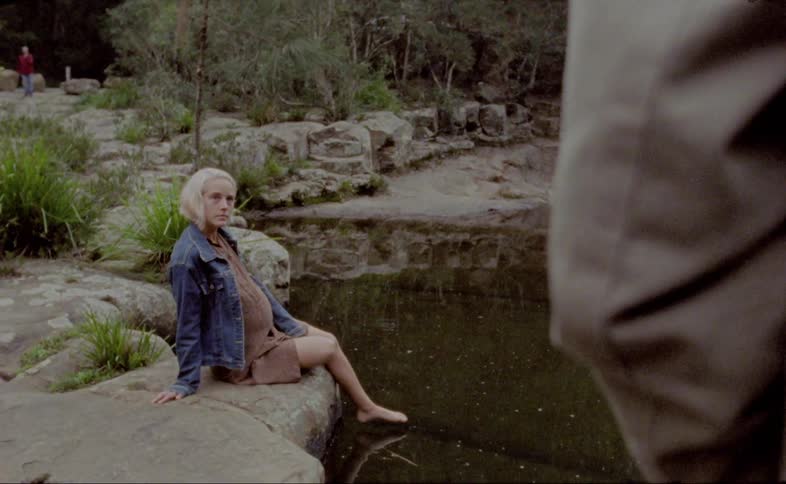 A pregnant blonde female sitting near a river with her foot in the water