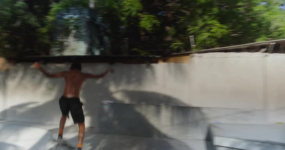 A young man with no shirt is wearing shorts as he hops on a skateboard and begins skateboarding and doing tricks around a skate park