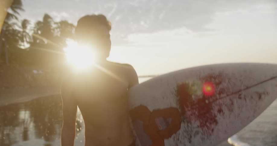 A man walking on the beach with a surfboard with the sky and the sun in the background