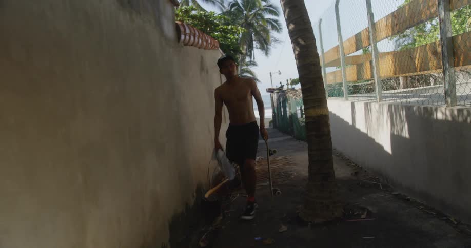 A young shirtless hispanic man carrying a skateboard through an alleyway near the beach