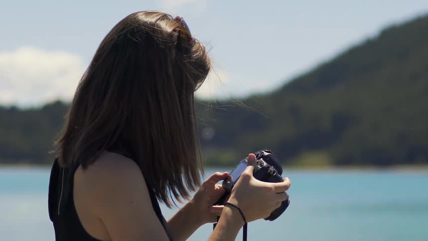 A woman standing near a body of water takes a picture then reviews it then sets to take another picture