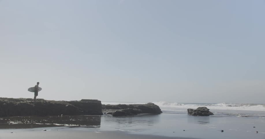 A man is standing on some rocks as he holds his surfboard and watches the waves crash into the beach