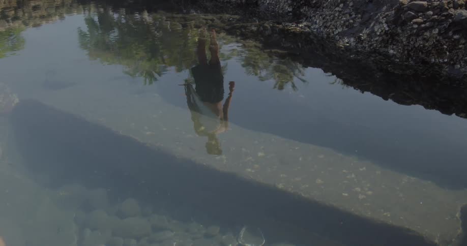 Man holds a surfboard as he looks at his reflection in the water