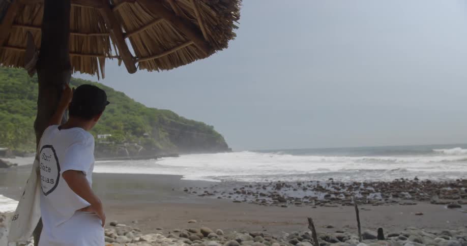 A man stands beside a tiki hut looking to the ocean water