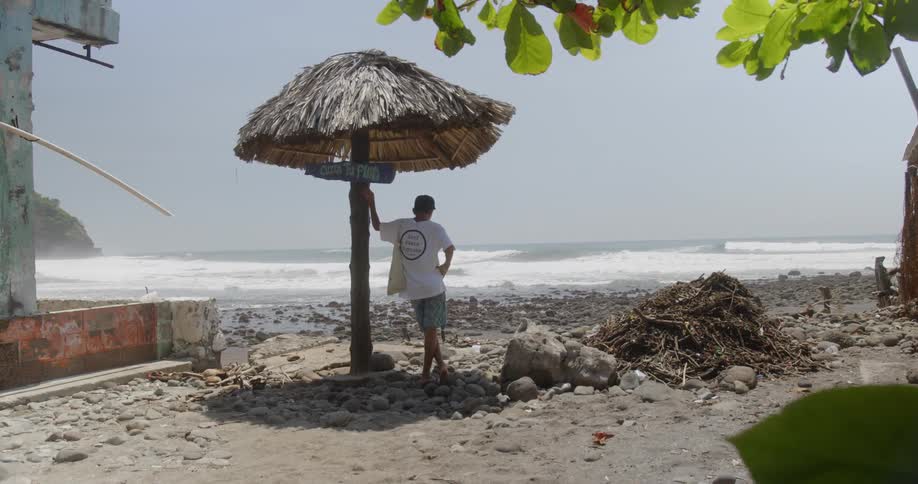 Male with hands on waist legs crossed standing under straw umbrella looking out at the beach