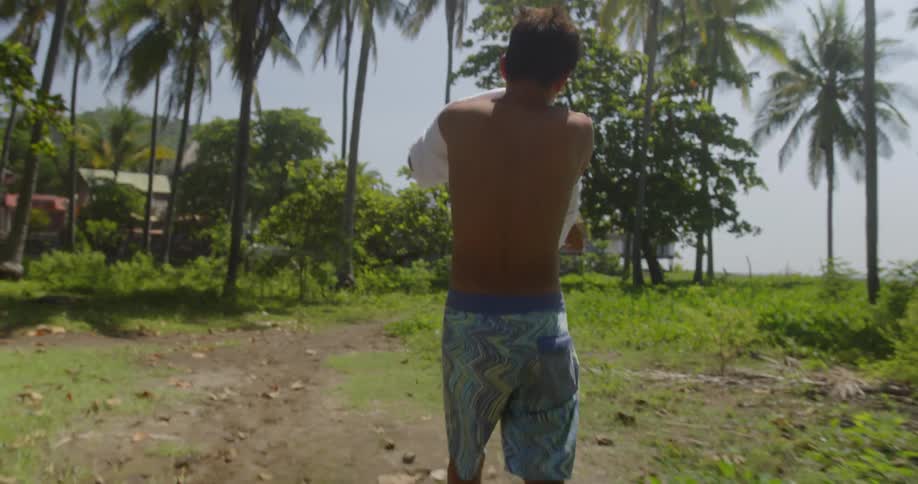A young male wearing board shorts walks along a dirt path amidst several palm trees in a tropical area as he puts on a white teeshirt