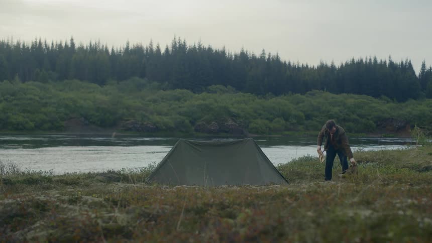 A man folds a towel and picks up a backpack beside a tent and lake