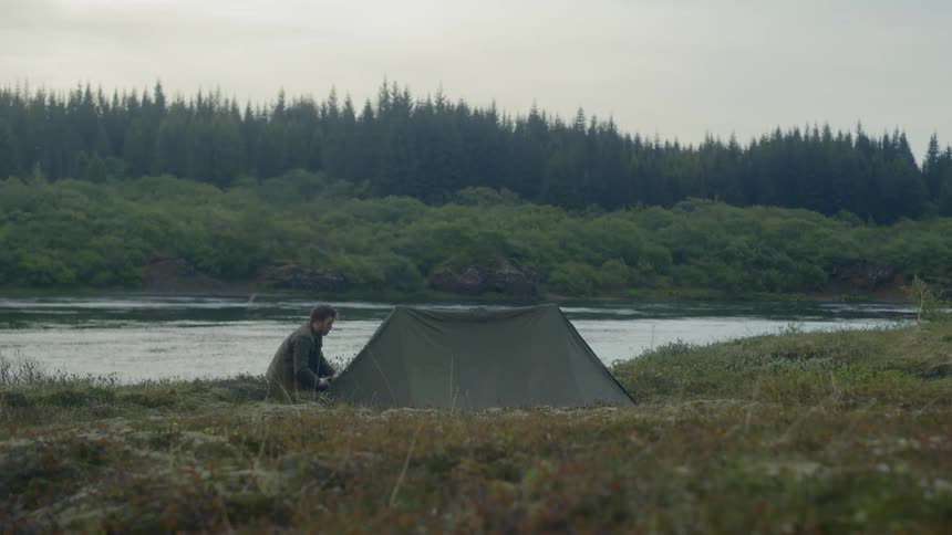 A man sets up a tent on the edge of a river