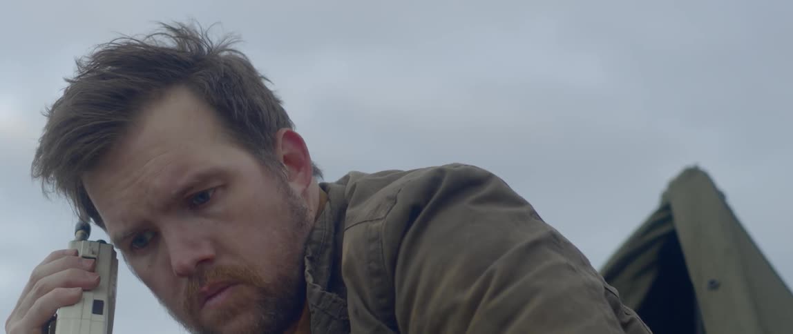 A man holds a walkie talkie up to his ear while crouched down outside in front of a tent