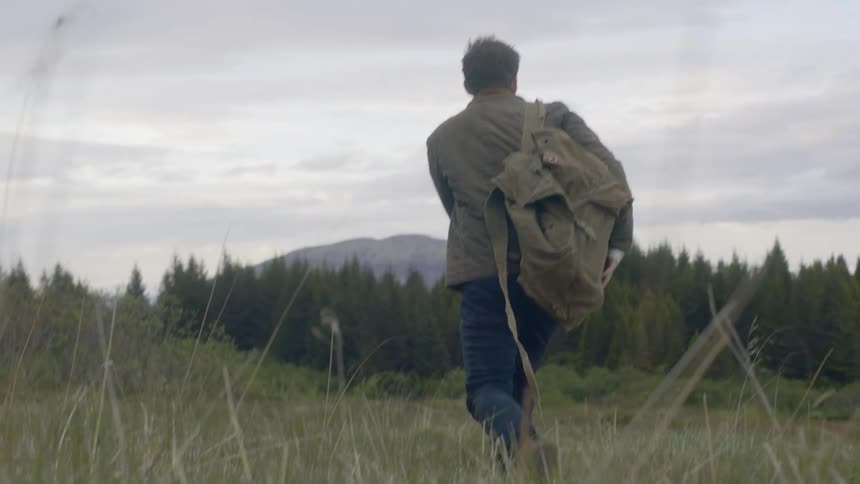 A hiker walks across a grassy field with a backpack near pine trees