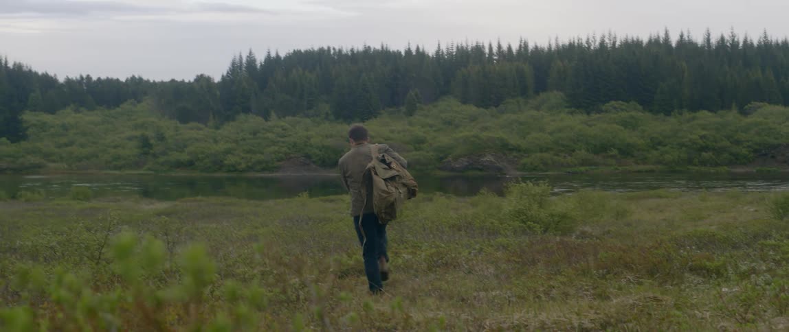A man carrying a backpack walks across a field towards a lake