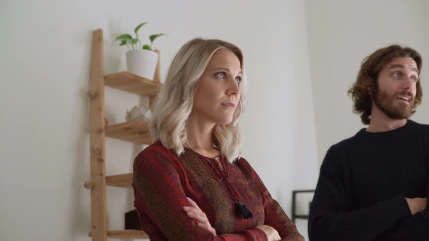 A man and a woman stand in a room with their arms crossed and looking around