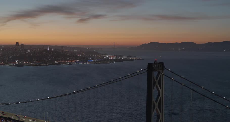 A suspension bridge is illuminated by lights as the sun sets over a nearby city