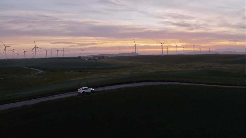 A white car travels a deserted road past wind turbines