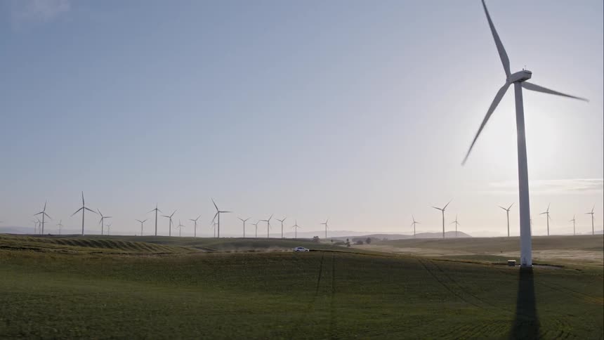 A car traveling on a dirt road leaves a dust trail as it pass fields and wind turbines