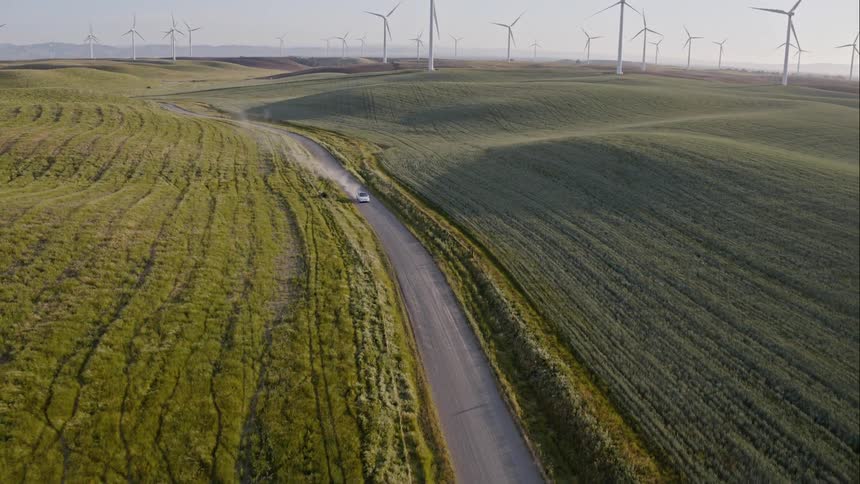 A car travels on a rural road past crop fields with wind turbines on the horizon