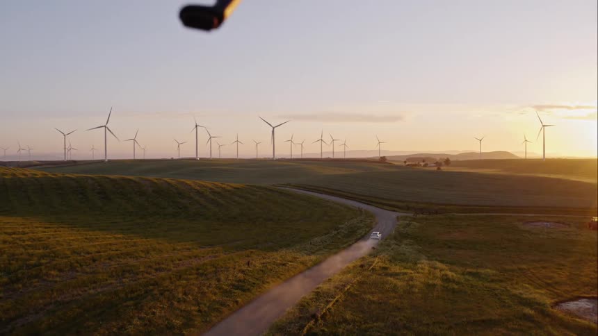 An aerial view of a white car driving on a rural road near a wind farm at sunset