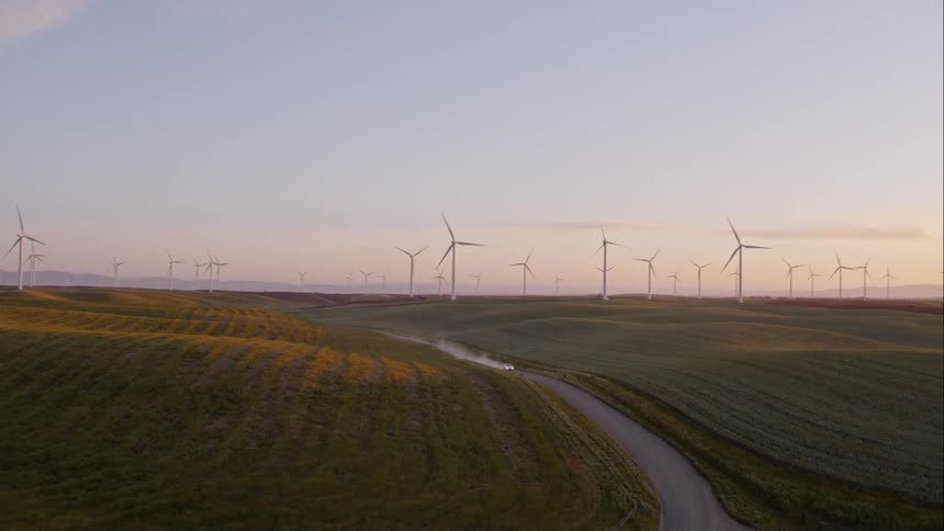 An aerial view of a car driving on a dirt road near a wind farm at sunset