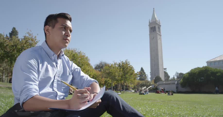 A man sits on the green park grass writing with a pencil on note paper pauses to look around
