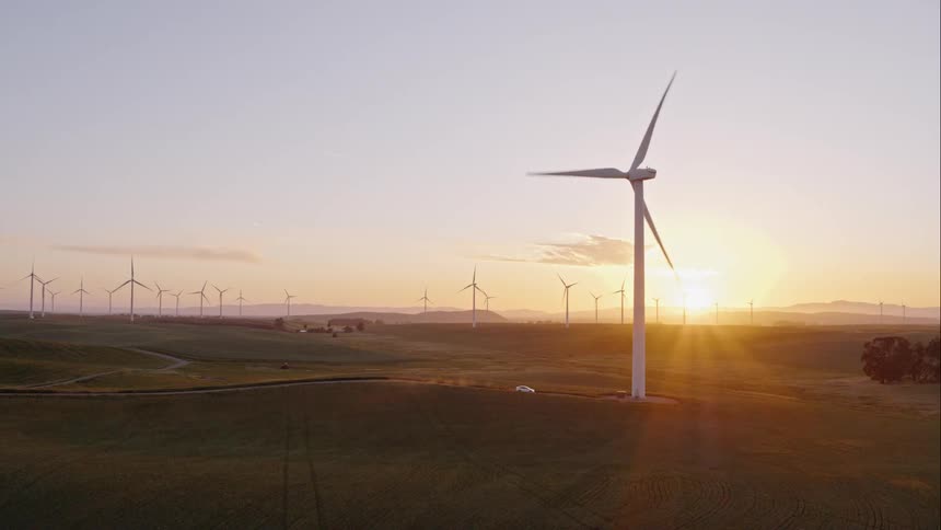 A car travels on a dirt road past wind turbines at sunset