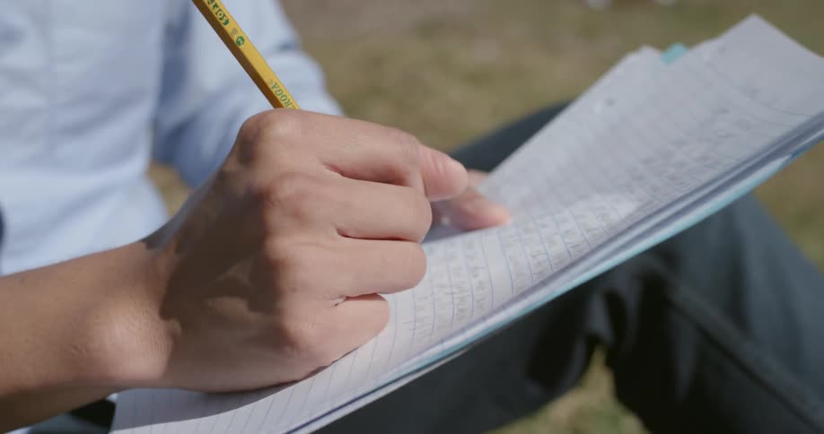 A man writes with a pencil on a piece of paper while sitting in the grass