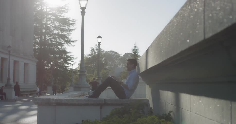 A man reads a book while leaning against a stone wall outside at a university