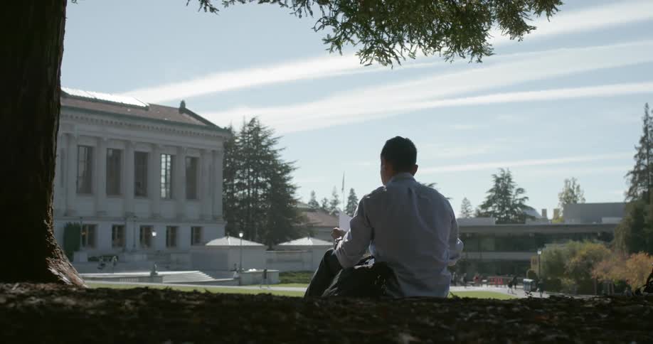 A man sits under the shade of a tree and reads from note paper hes holding