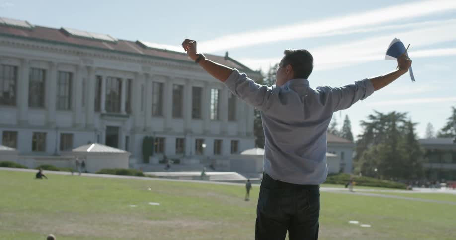 A man holding notepaper and a pencils stretches while standing on a lawn near a large building