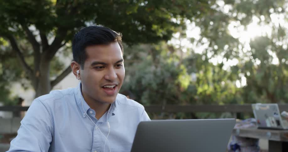 A young man in a blue button up shirt is talking to someone through his laptop