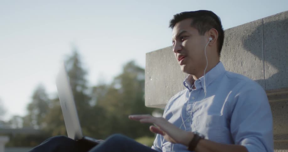 A man wearing headphones smiles while video chatting on a computer outside