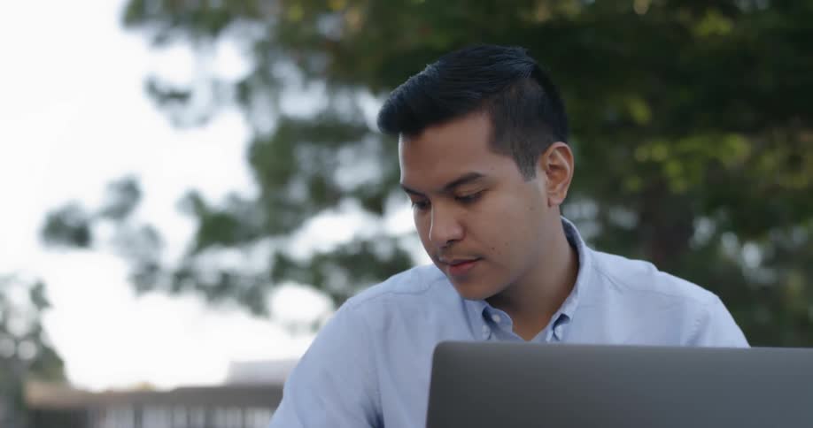 A young man sits and writes on a paper with his laptop open in front of him