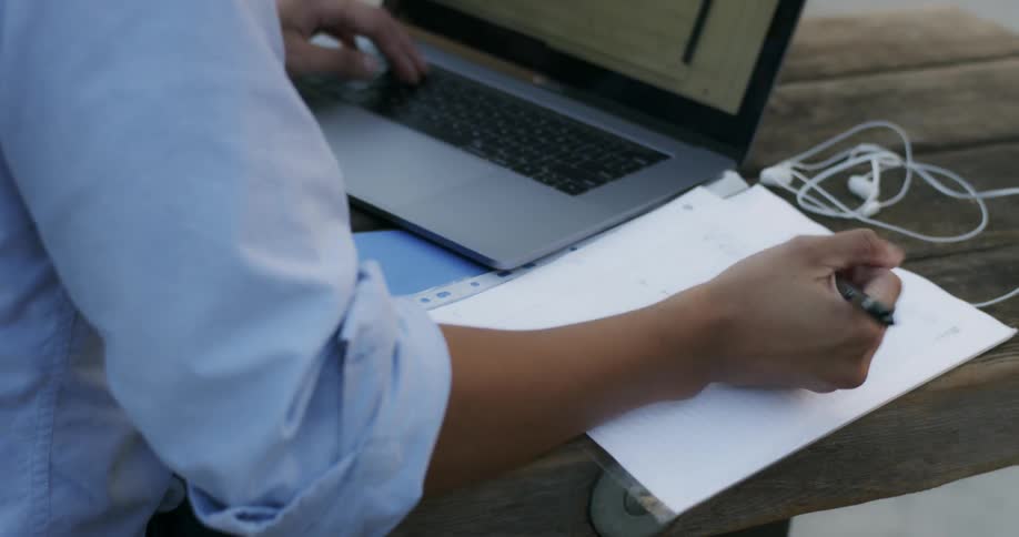 A person sitting at a table using a touchpad on a laptop and writing on a sheet of paper